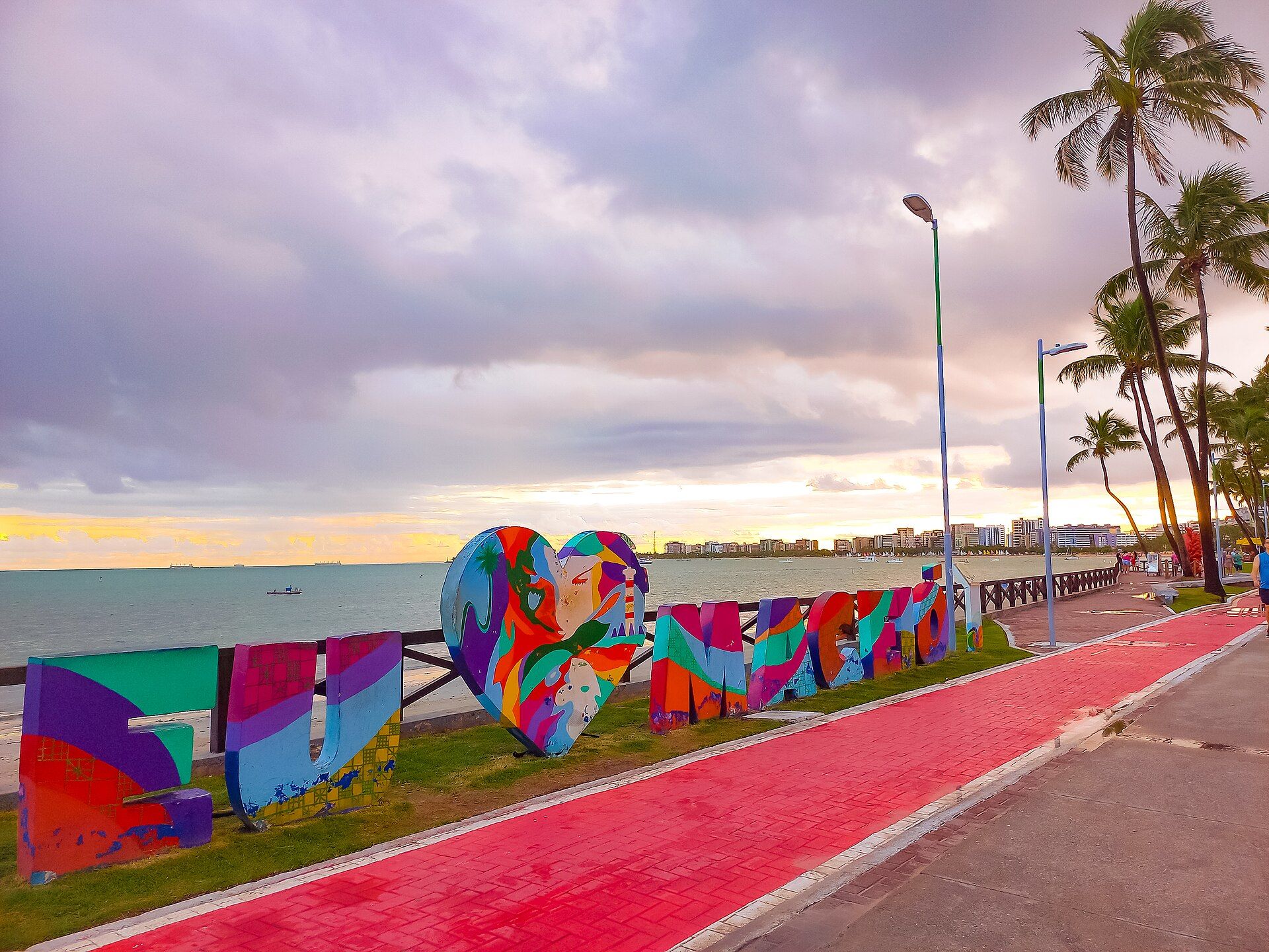 Letreiro colorido EU ♥ MACEIÓ na orla da Pajuçara, com letras pintadas em turquesa, roxo, vermelho e laranja e o mar ao fundo no fim de tarde