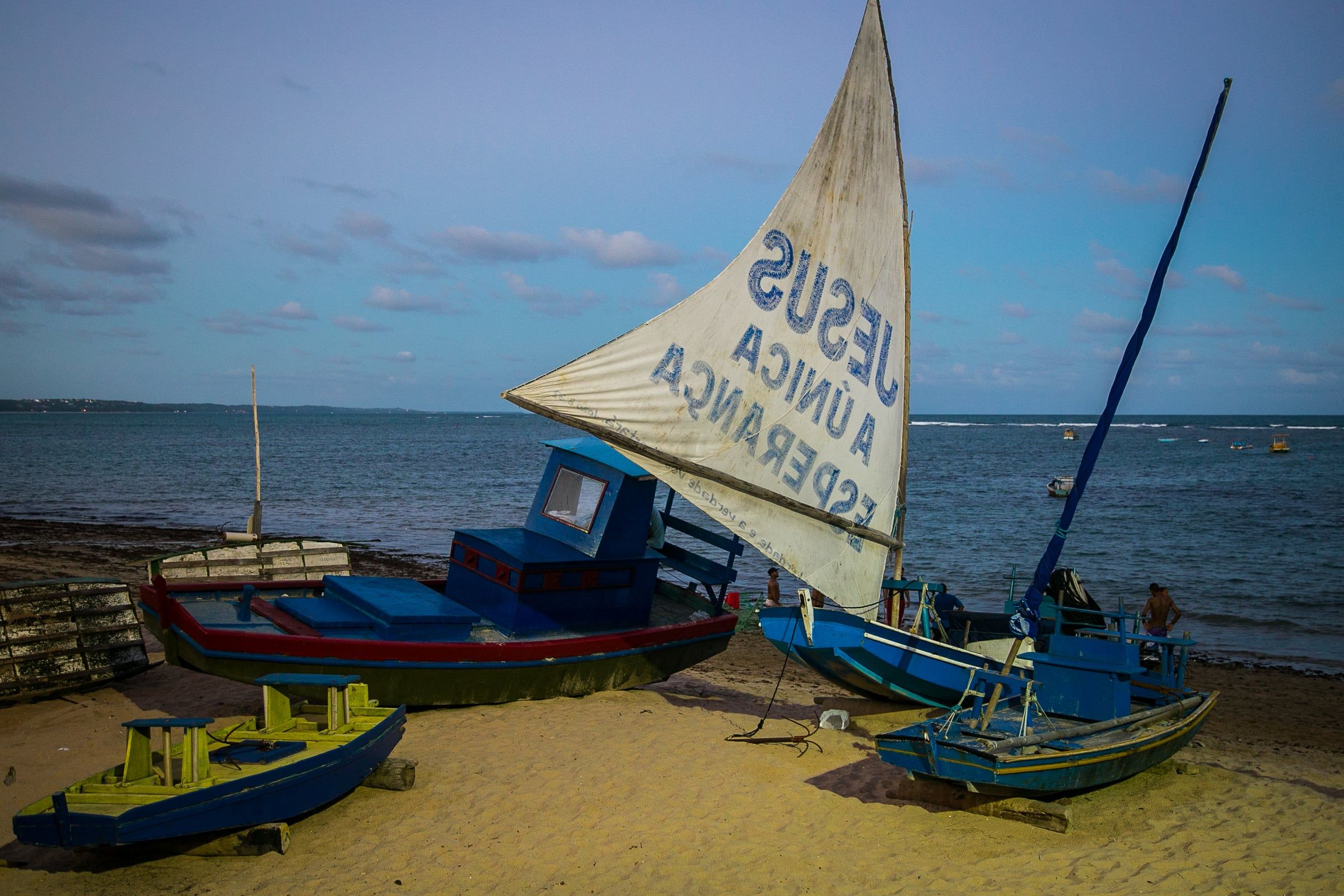 Jangada de vela branca e barcos coloridos na areia da Praia de Jatiúca